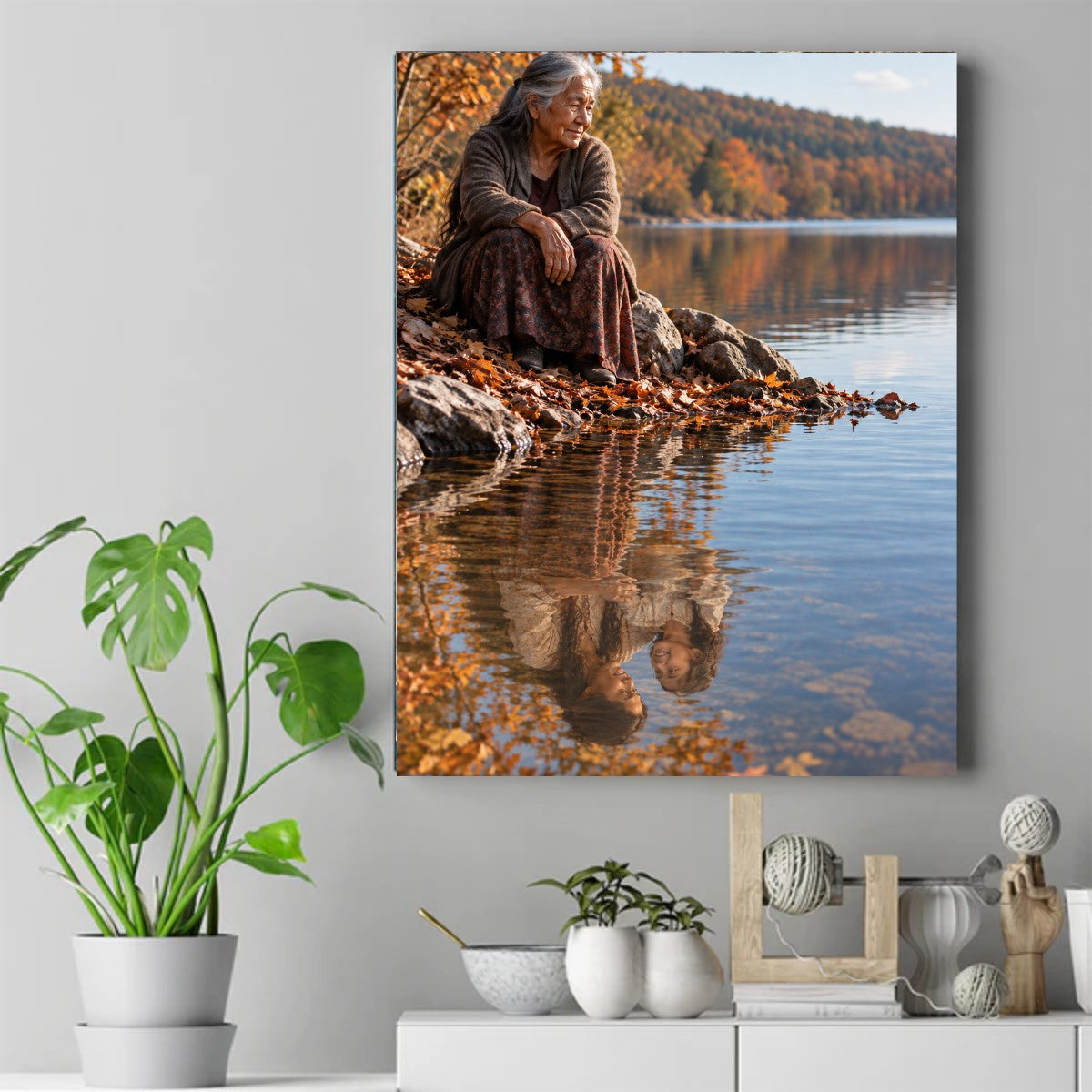 Elderly Native woman in brown cardigan sitting on rocky shore by autumn lake, her face reflected in calm water. – ảnh 2